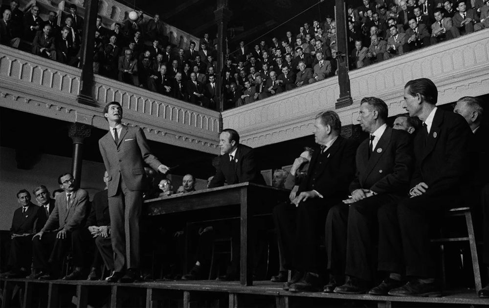 Still image from The Trial. Anthony Perkins as K stands on the edge of a stage surrounded by onlookers at his trial. 