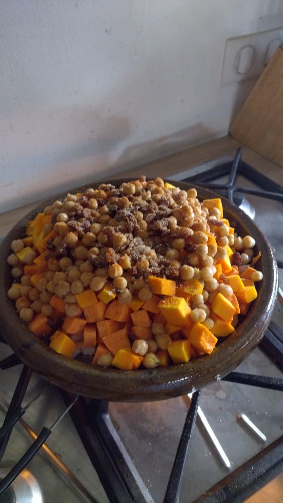 Un plat à tajine sur la plaque de cuisson à gaz avec des oignons, du céleri, des navets 
Tous ces ingrédients sont cachés par de la patate douce, butternut, des pois chiches et des raisins.
Il y a du poivre noir en grain, du gros sel, de la coriandre en poudre, de l'anis vert et un peu de piment doux fumé 