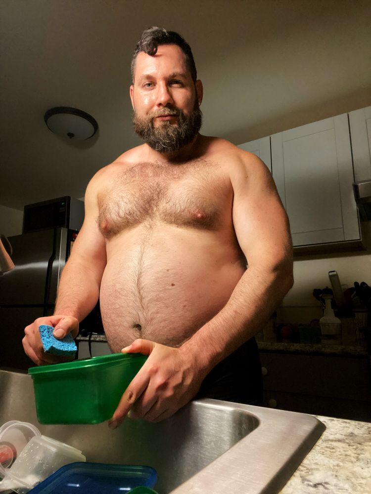 A sweaty, shirtless man in the kitchen doing dishes while flexing his muscles and showing off his big, fat belly.