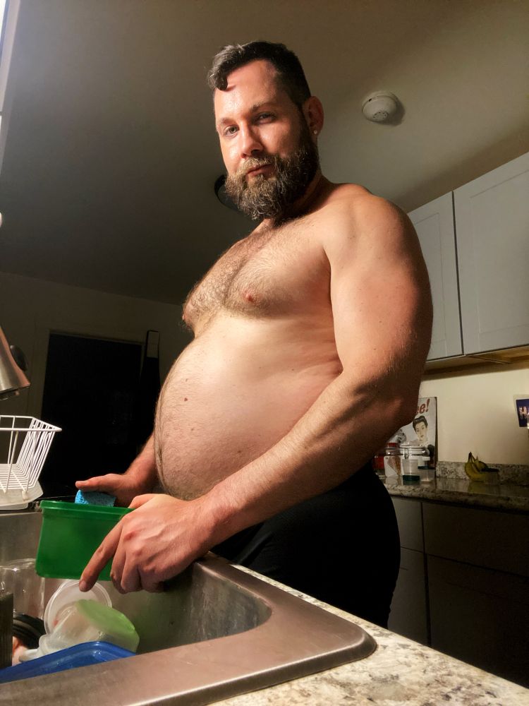 Side view of a shirtless man in the kitchen doing the dishes while flexing his muscles and showing off his big, fat muscle belly after a big post-workout meal.