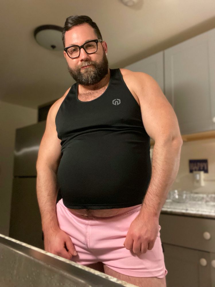 Man in tight gym clothes showing off his muscles and big fat belly after eating a big post-workout meal. His swollen lower belly is popping out of the bottom of his shirt and pushing his waistband down.