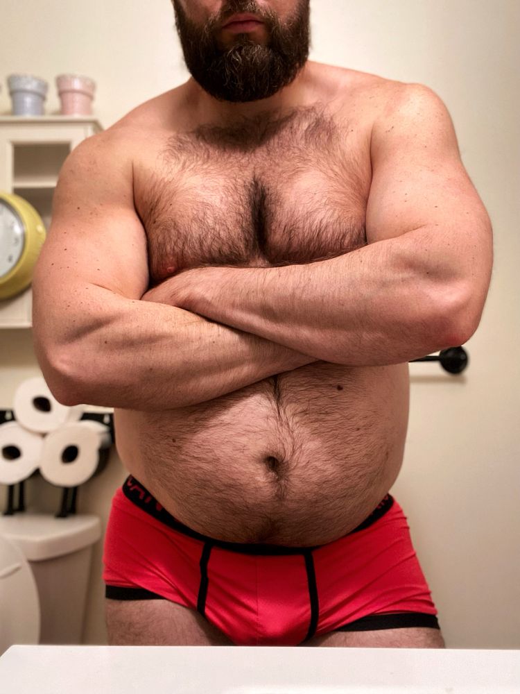 Shirtless man flexing his muscles while crossing his arms over his big hairy belly in pink underwear.