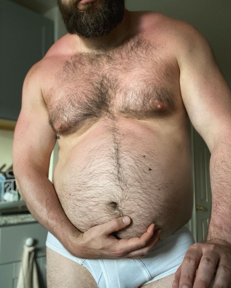 Man with sunburn holding his big fat belly while flexing his muscles in white underwear.