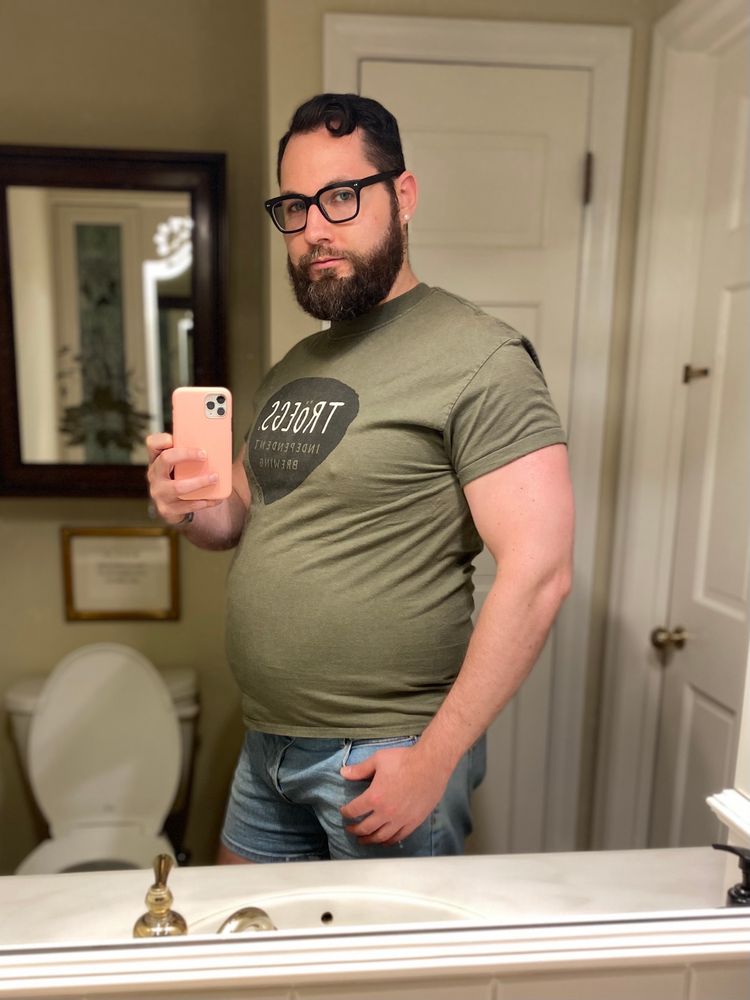 Man posing in a bathroom mirror wearing a tight green shirt and flexing his muscles while showing off his big belly.