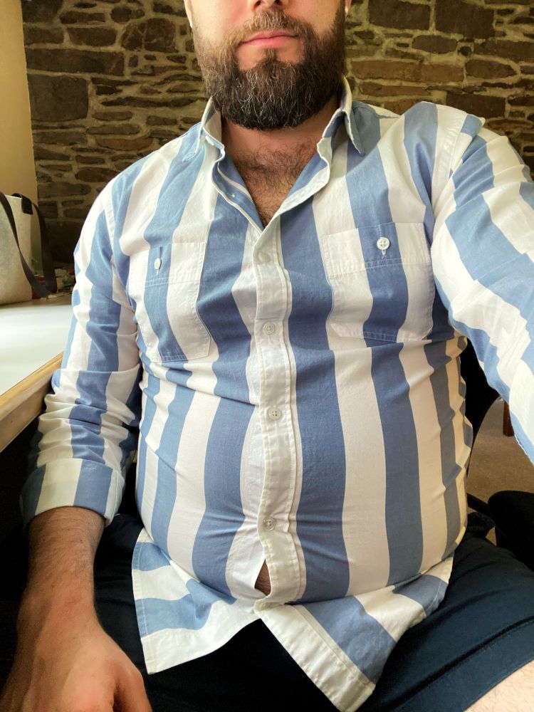 Man in a blue and white striped button down shirt that is too small and is exposing his big belly.