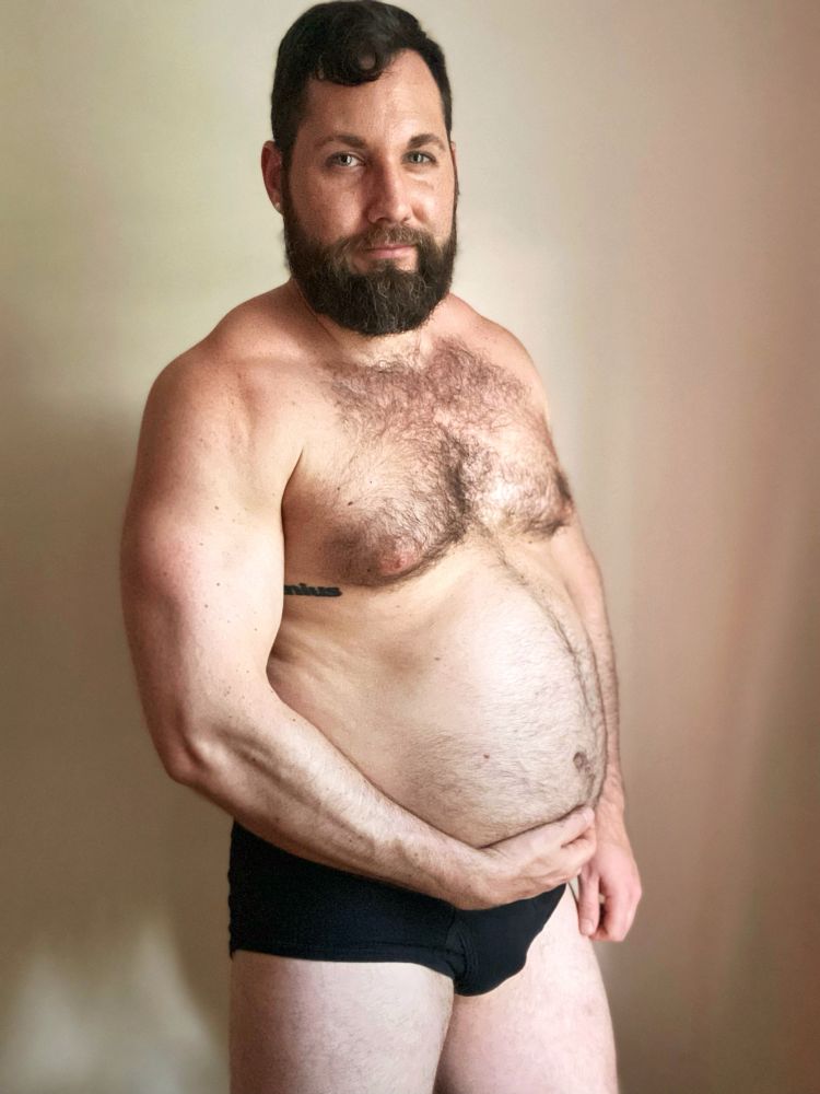 Man standing in a side pose in black underwear while flexing his bicep and holding his big stuffed belly.