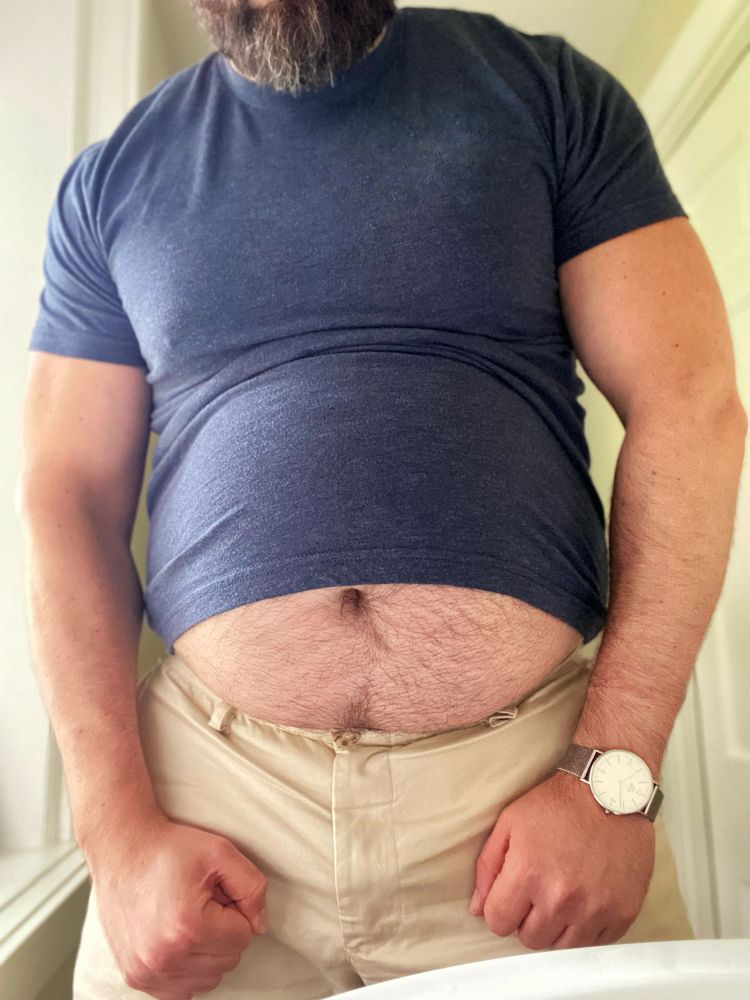 Man flexing his muscles while wearing a tight blue shirt that’s pulled up to show off his big fat belly.