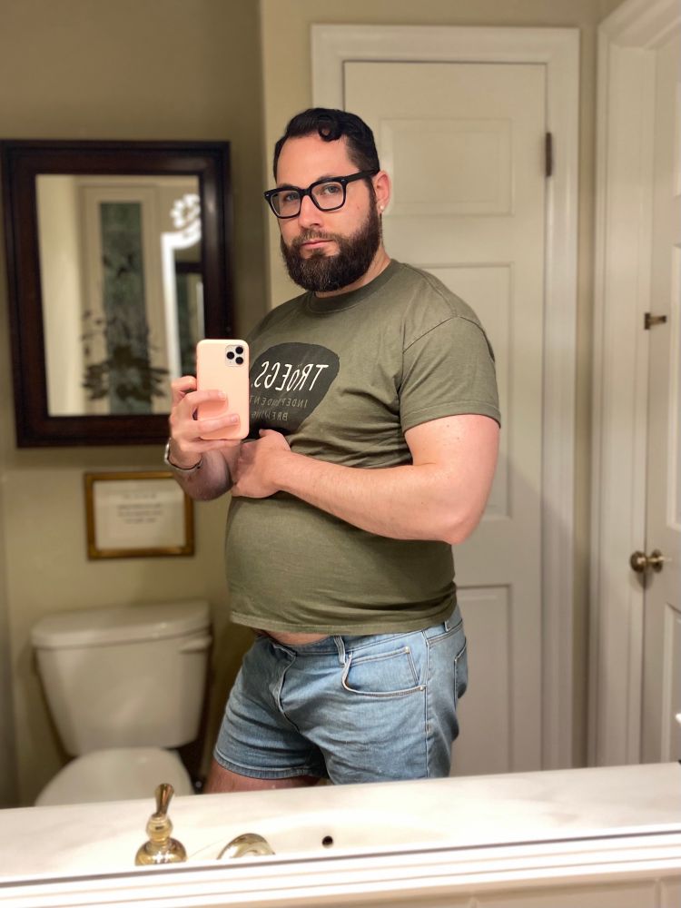 Man posing in a bathroom mirror wearing a tight green shirt, flexing his muscles, and showing off his big belly as it sticks out from the bottom of the shirt.