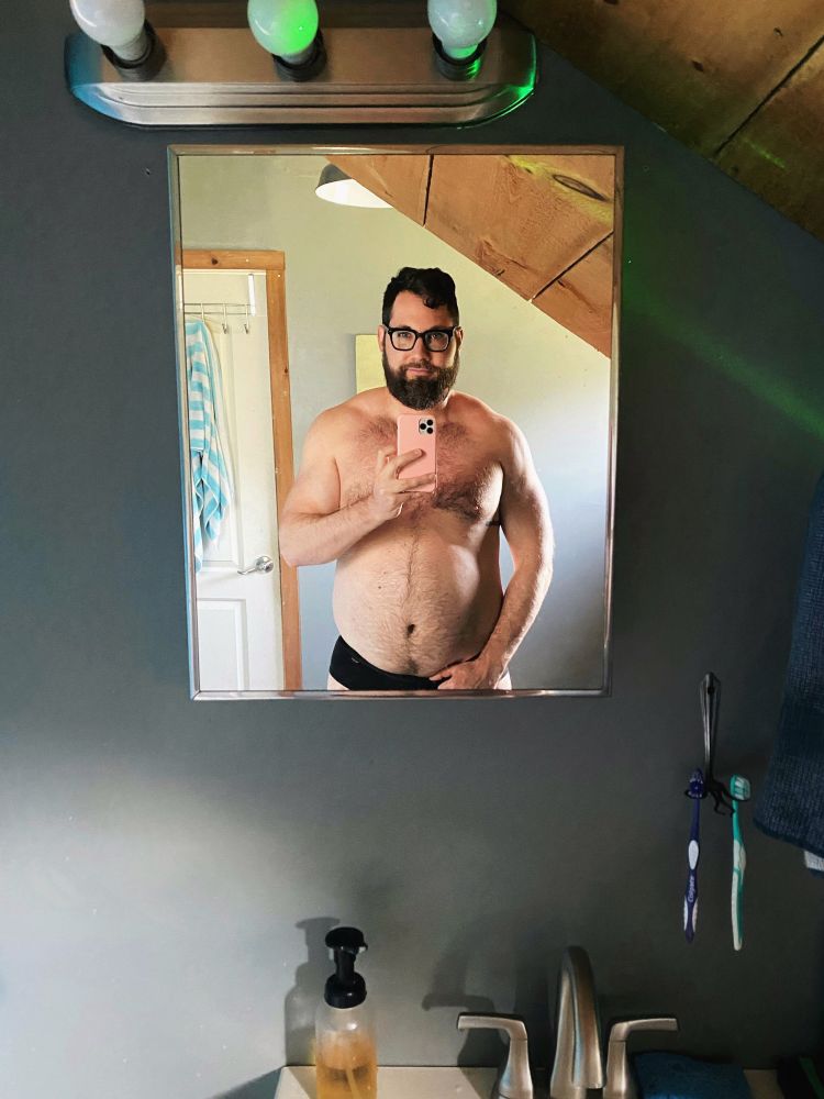 Beefy man taking a selfie in a bathroom mirror while shirtless and flexing his muscles wearing black underwear.