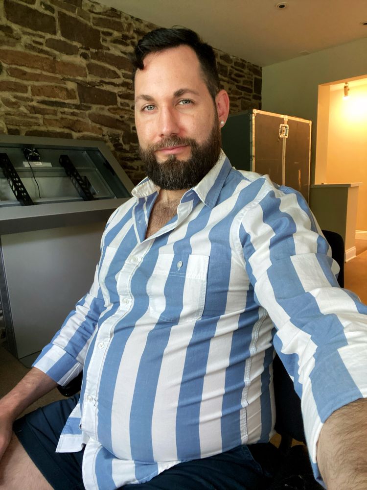Man in a blue and white striped button-down shirt that is too small and is tight across his big belly.