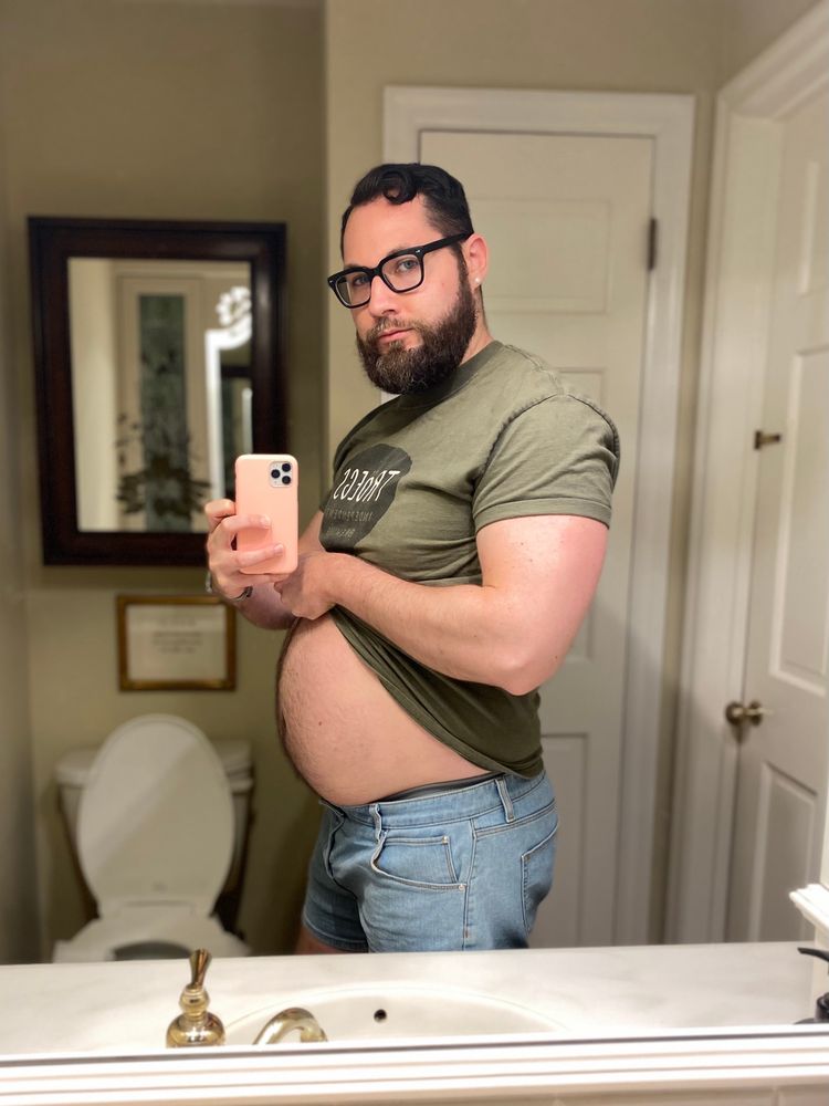 Man posing in a bathroom mirror wearing a tight green shirt and flexing his muscles with the shirt pulled up to show off his big belly.