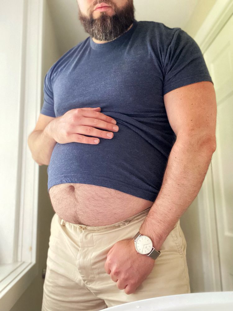 Man posing with his big fat belly out while wearing a tight blue shirt and flexing his beefy muscles.