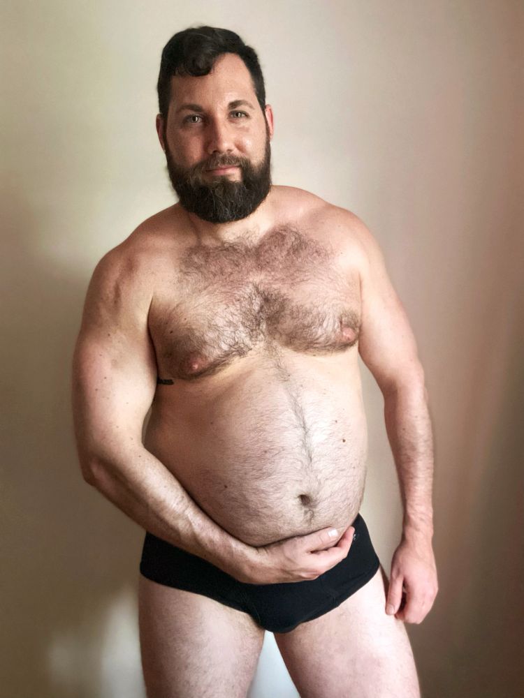 Man posing in black underwear while flexing his muscles and holding his big stuffed belly.
