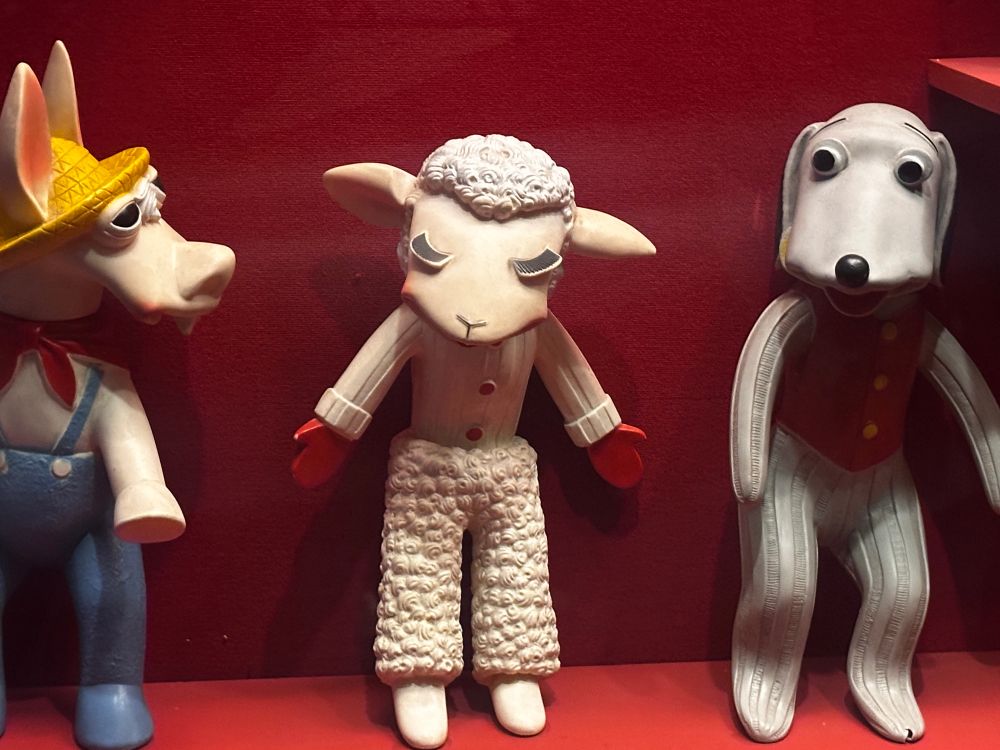 A photo of the puppet lamb chop between the other characters of the show including a dog and a donkey. They look upon her, judging. While she looks exasperated do to the terrible overhead lighting  