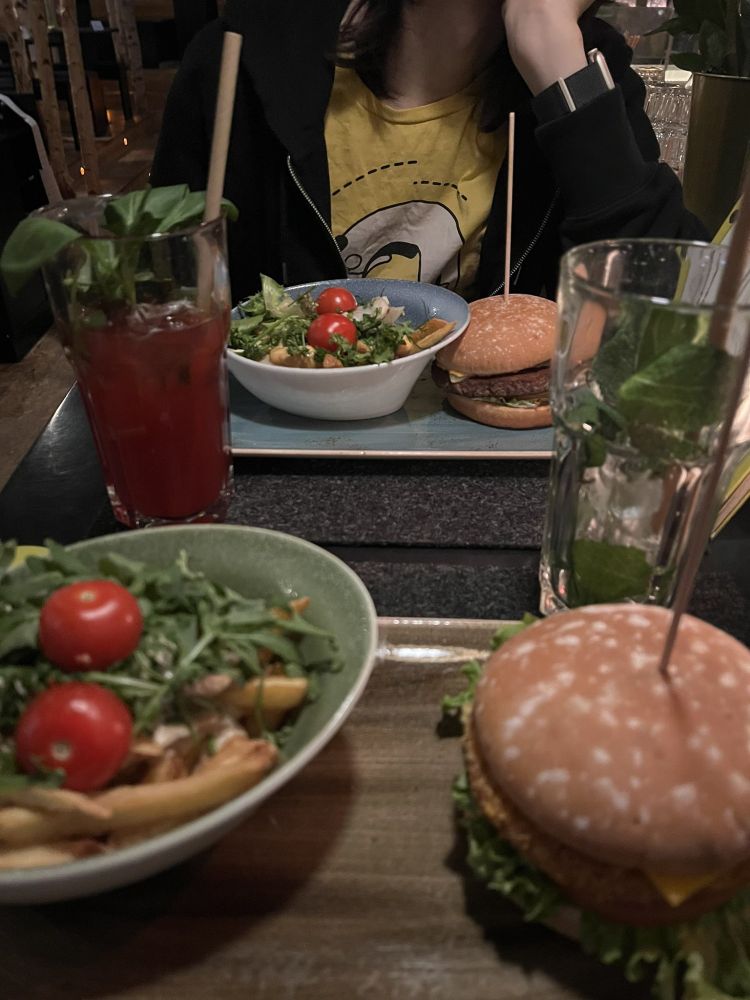 A dimly lit restaurant table with two meals. In the foreground, there is a burger with lettuce and cheese on a wooden plate, alongside a green bowl containing salad with cherry tomatoes and arugula on top of fries. In the background, a second plate features a similar setup with a burger and a salad. Two beverages are present: a red drink garnished with basil and a clear drink with mint leaves. A person in a yellow kagi shirt and black jacket is seated behind the second plate.
