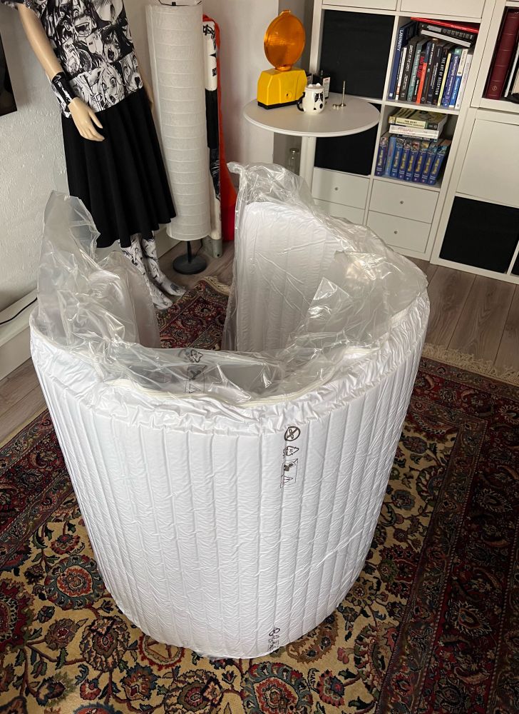 A rolled-up white mattress, partially wrapped in clear plastic packaging, is placed on a patterned rug in a room. The background features a mannequin wearing a black skirt and a top with a graphic design, a tall cylindrical lamp, a small round table with items on it, and a bookshelf filled with books and other objects.