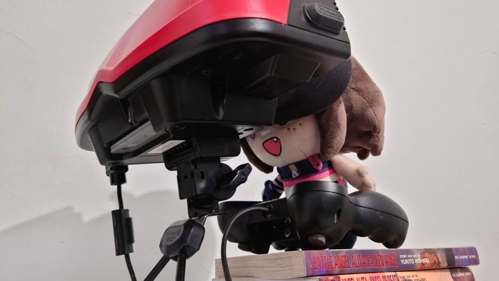 Camera has moved to show it's an Ascari plush with her head shoved in the Virtual Boy, standing on a stack of "Battle Angel Alita" manga volumes