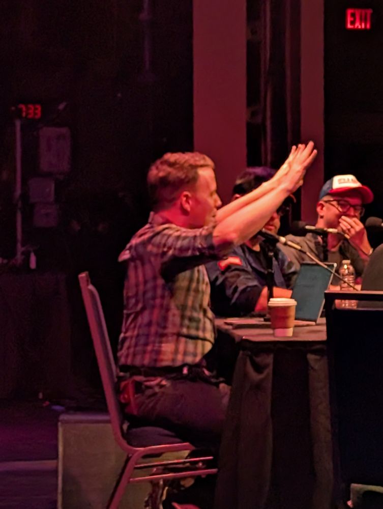 Brennan Lee Mulligan gesturing as he narrates, and beyond him Griffin is clearly smiling while holding on a false moistache