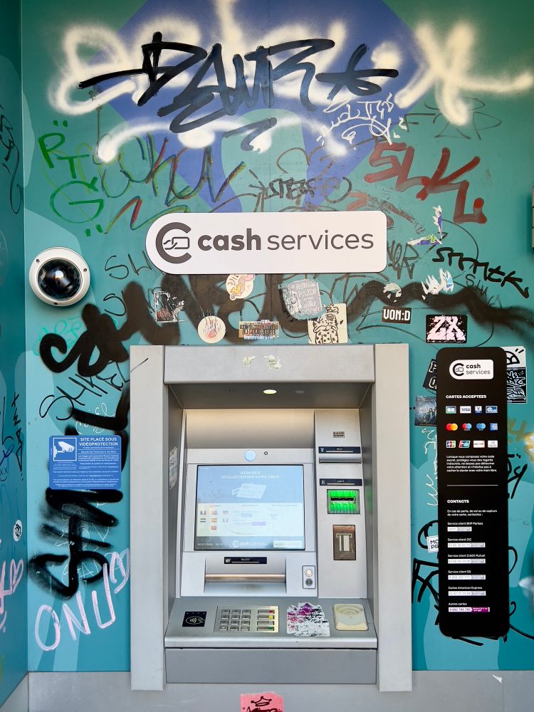 Das Bild zeigt einen Geldautomaten, der von zahlreichen Graffitis und Kritzeleien umgeben ist. Es vermittelt die Botschaft eines urbanen, alltäglichen Szenarios, das von Kunst und Vandalismus zugleich geprägt ist. Möglicherweise drückt der Künstler eine Verbindung zwischen dem nüchternen Zweck eines Geldautomaten und der lebhaften, chaotischen Energie der Stadt aus. Die bunte und vielförmige Gestaltung der Wand könnte darauf hindeuten, dass der Künstler Freiheit und Kreativität schätzt, während die Präsenz des Überwachungskamerasymbols vielleicht auf eine kritische Haltung gegenüber moderner Überwachungsgesellschaft hinweist. Die Emotionen könnten zwischen ausgelassener Freude und einem subversiven Gefühl der Rebellion schwanken, was in der jugendhaften Ausdrucksweise der Graffitis widergespiegelt wird.