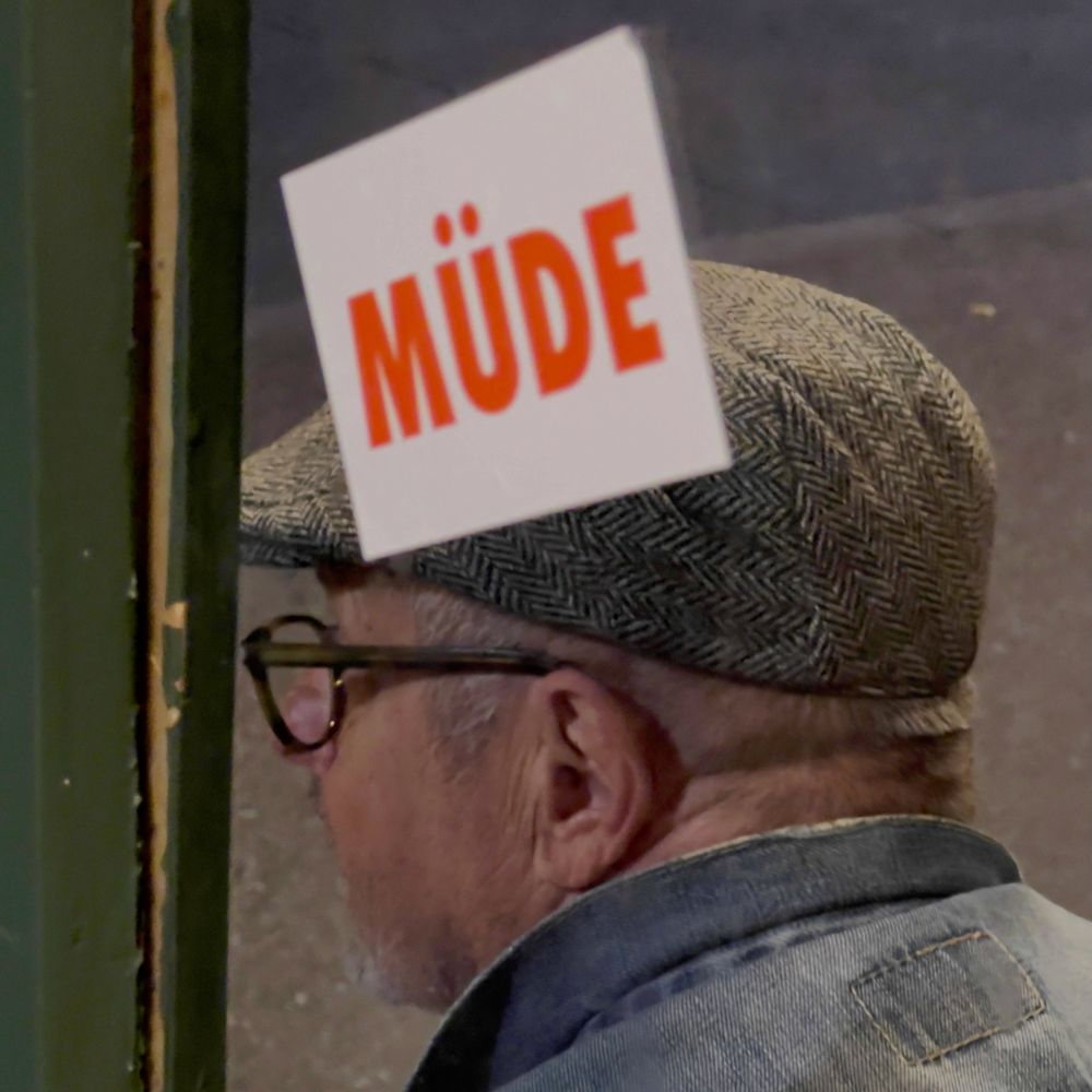 Das Bild zeigt einen älteren Mann mit einem Schiebermützähnlichen Hut und Brille, der zur Seite blickt. Das Wort 'MÜDE' ist in auffälligen roten Buchstaben auf einem Schild oder Aufkleber zu sehen, das offenbar unmittelbar vor ihm platziert ist. Die Botschaft 'MÜDE' könnte symbolisch für eine allgemeine Erschöpfung oder Müdigkeit stehen, sei es körperlich oder emotional. Die Position des Schildes und die Haltung des Mannes erweckt den Eindruck von Nachdenklichkeit oder Rückzug. Die nach hinten geneigte Körperhaltung des Mannes und das leicht unscharfe Umfeld deuten auf eine ruhige, isolierte Atmosphäre hin, die diese Müdigkeit unterstreicht. Es könnte ebenfalls ein Ausdruck von Selbstreflexion oder Melancholie sein, was tiefere emotionale Schichten in dieser einfachen, aber ausdrucksstarken Bildkomposition andeutet.