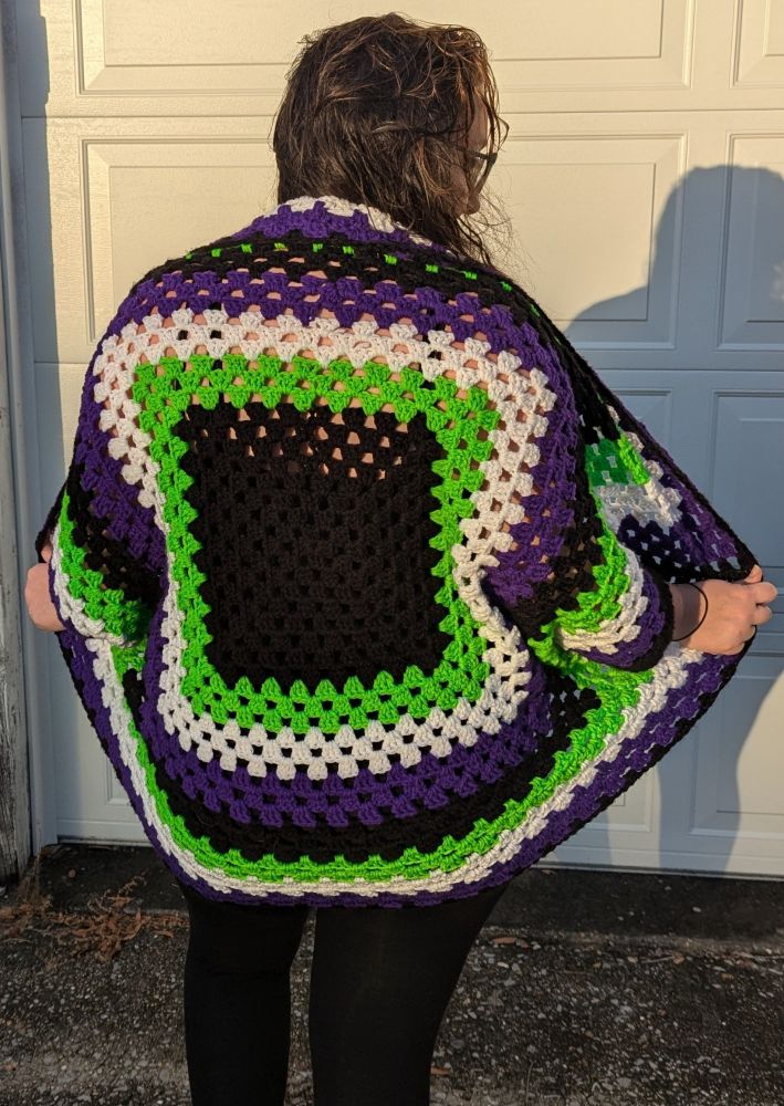 Meg standing in front of a white wall, wearing black leggings and a crocheted sweater.
The sweater is done with granny stitches in black, green, white, and purple square rounds.
