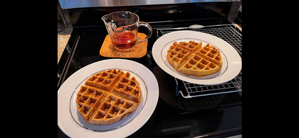 Two Lemon-Poppy Seed waffles  on plates with a 2c liquid measuring cup filled with warmed maple syrup on a glass top stove. 