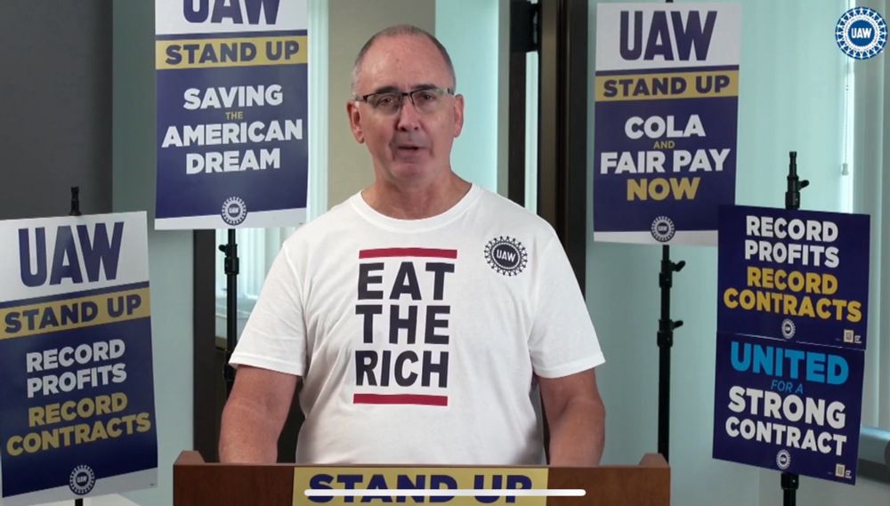UAW President Shawn Fain standing at a podium wearing a shirt that says eat the rich.