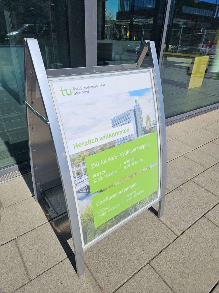 Ein Aufsteller vor dem Eingang des ITMC in Dortmund mit einem Poster, auf dem "Herzlich Willkommen" steht, und zu den beiden Tagungen ZKI AK Web und Confluence.campus eingeladen wird.
