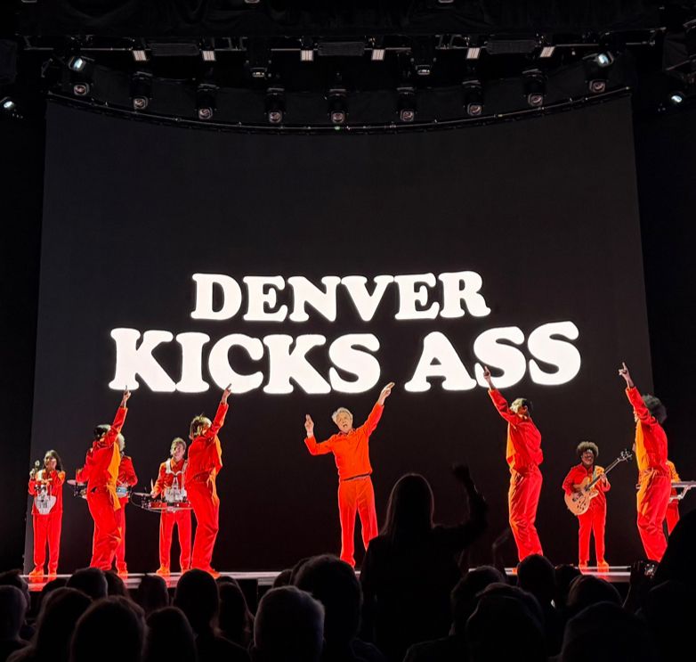 David Byrne surround by band members all dressed in red. “Denver Kicks Ass” is projected behind them in a white font with a black background.