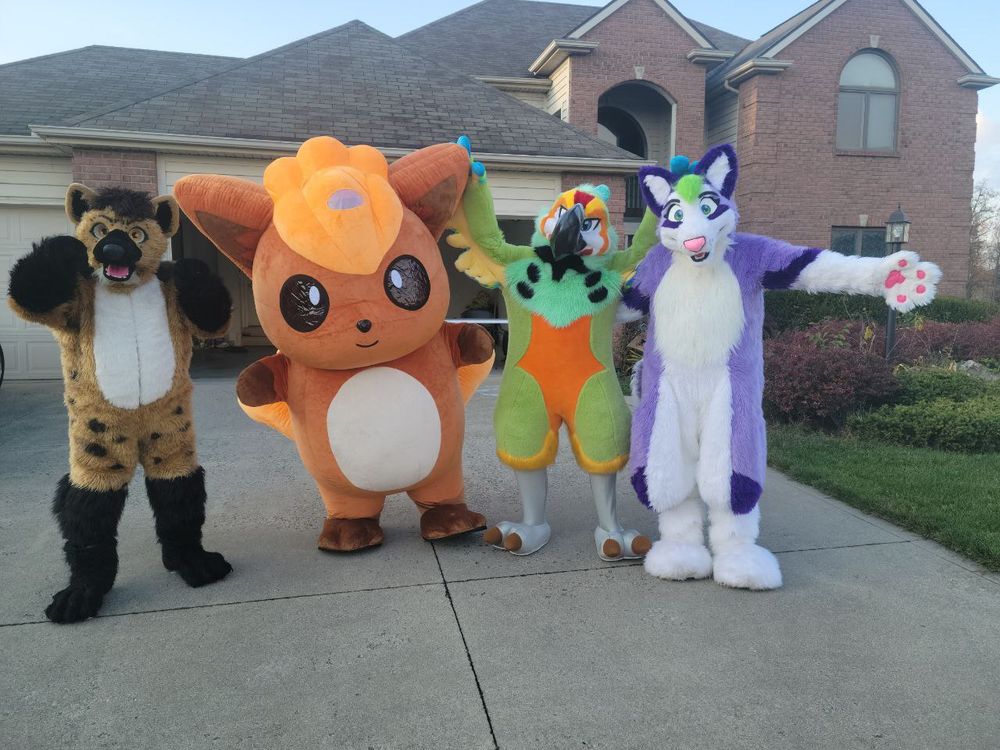 Four fursuits in a row, in a driveway, with a fancy tan house behind. Starting from the left is Savannah, a tan spotted hyena with black hair, snout, arms and legs, and a white belly. Next is Vulpix, a large inflatable pokemon, orange with brown paws, white belly, and bright orange hair. Third is Capri, a harlequin macaw, a green bird with orange tummy, grey legs and feet claws, yellow inside her wings, with yellow, red and white markings on her face. Last is Susser, a purple husky with green hair, pink paw pads, and white muzzle, belly, arms and legs.