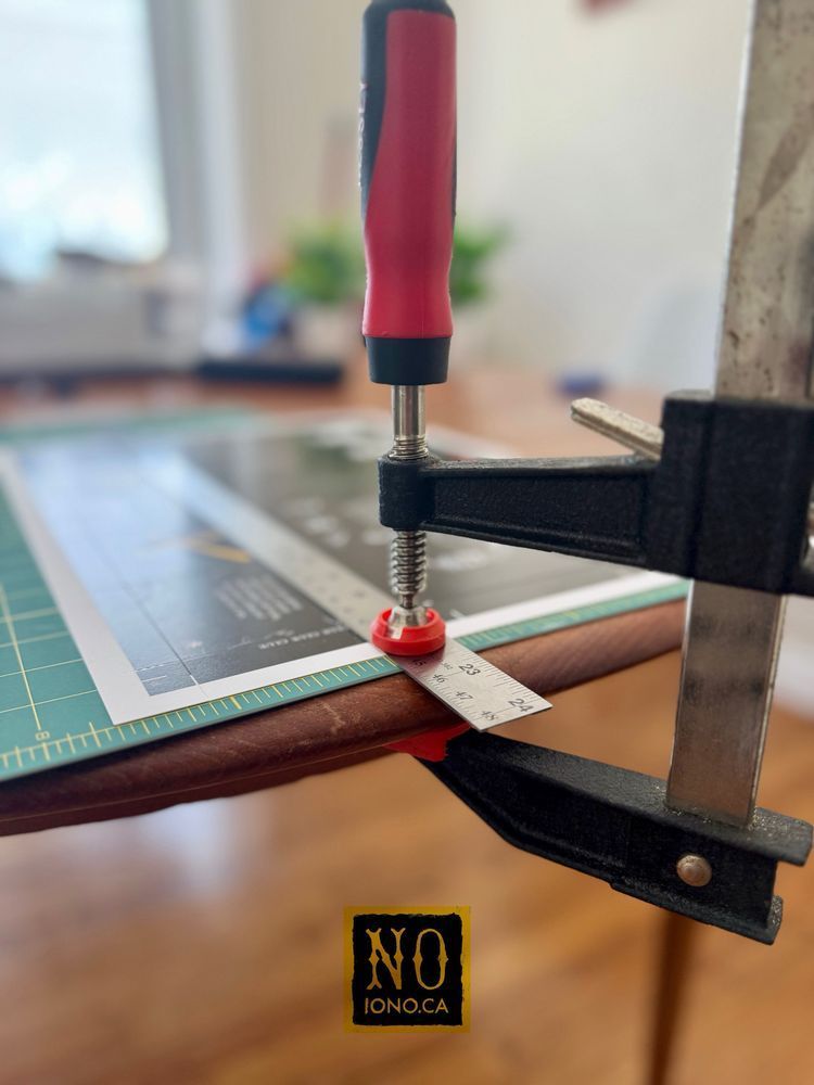 A close-up photo shows a red-handled clamp securing a metal ruler to the edge of a wooden table. A green self-healing cutting mat and printed material are positioned underneath the ruler, suggesting a setup for cutting or scoring paper accurately. The background is softly blurred, with natural light coming from a nearby window.