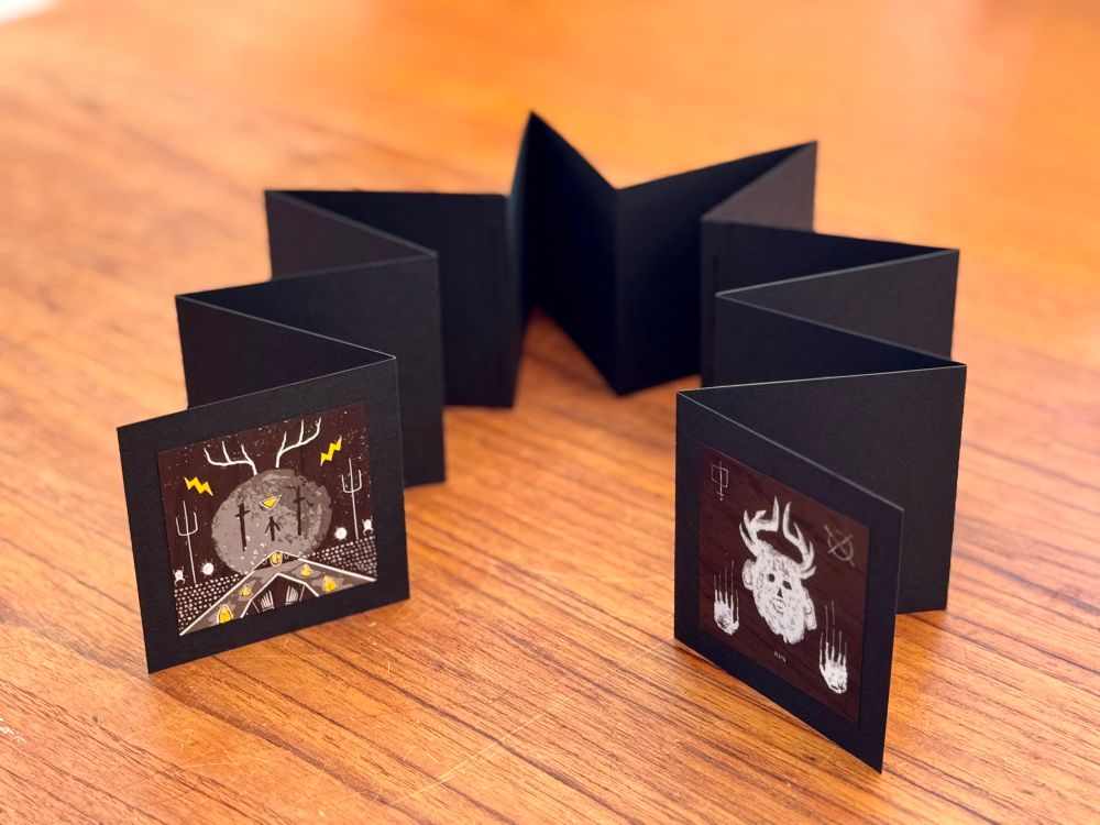 A photograph of a handmade accordion-style art book with black pages, opened and standing on a wooden surface. The panels display dark, surreal illustrations. On the left panel, a gray figure with antlers stands before a large moon-like circle, surrounded by yellow lightning bolts and trident-like symbols. On the right panel, a white, horned skull-like figure is framed by abstract glyphs and ghostly handprints. The tone is ominous and mythic, evoking a ritualistic or cultic atmosphere.