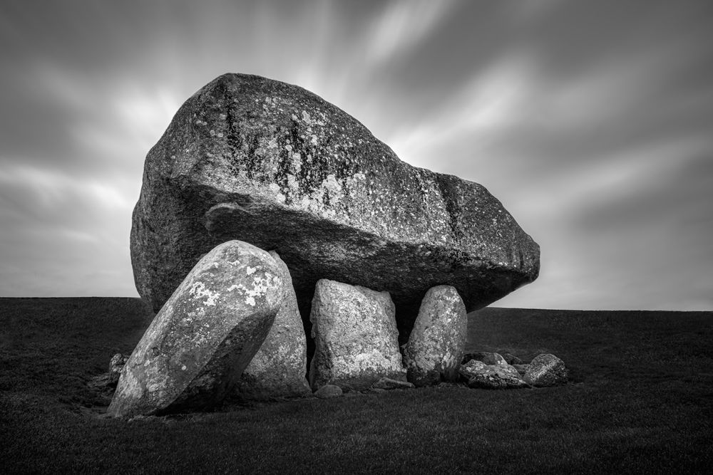 A black & white image of a 'portal tomb' (otherwise known as 'dolmen') found in County Carlow, Ireland. Constructed of a half dozen(??) or so standing stones with one large capstone resting atop those at a shallow angle. A long exposure technique to capture the movement of clouds across the sky and symbolize the passing of time for which these structures have endured. Such structures are fairly numerous in Ireland and believed to have been constructed between ~ 3800BC and 3000BC.