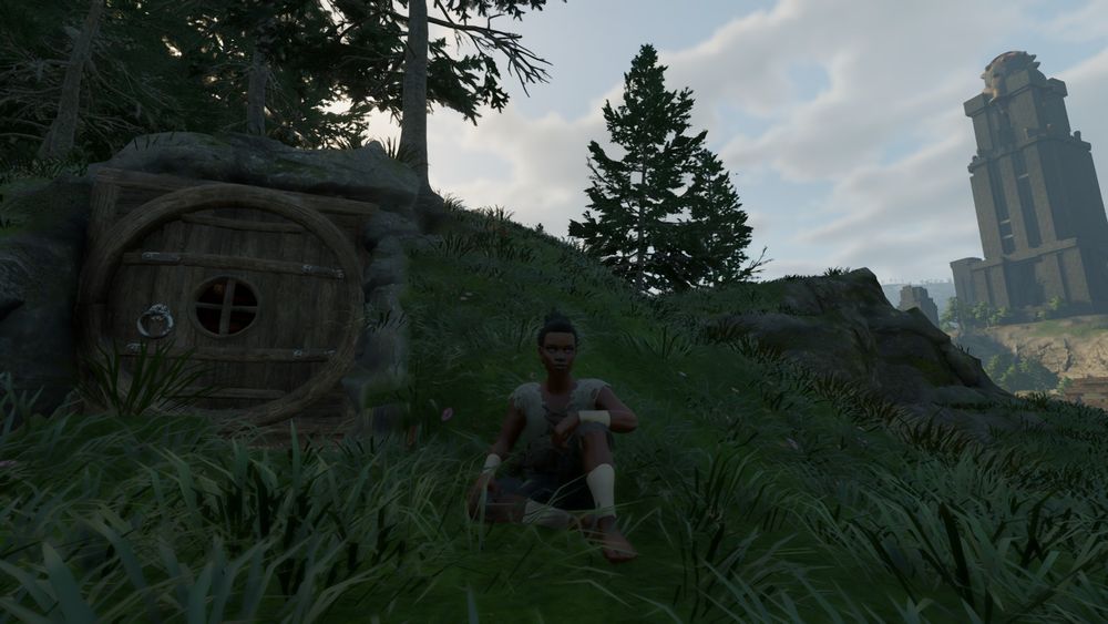 A small grassy hill with a circular wooden door leading into a small indoor space resembling a hobbit hole. In the background is a tall tower in the distance. Center frame is a woman sitting on the grass looking at the camera with a relaxed face.