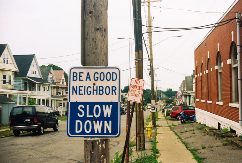 Film shot of a street and sidewalk. In the foreground are to street signs the first says BE A GOOD NEIGHBOR SLOW DOWN
the one behind it says NO PARKING HERE TO CORNER