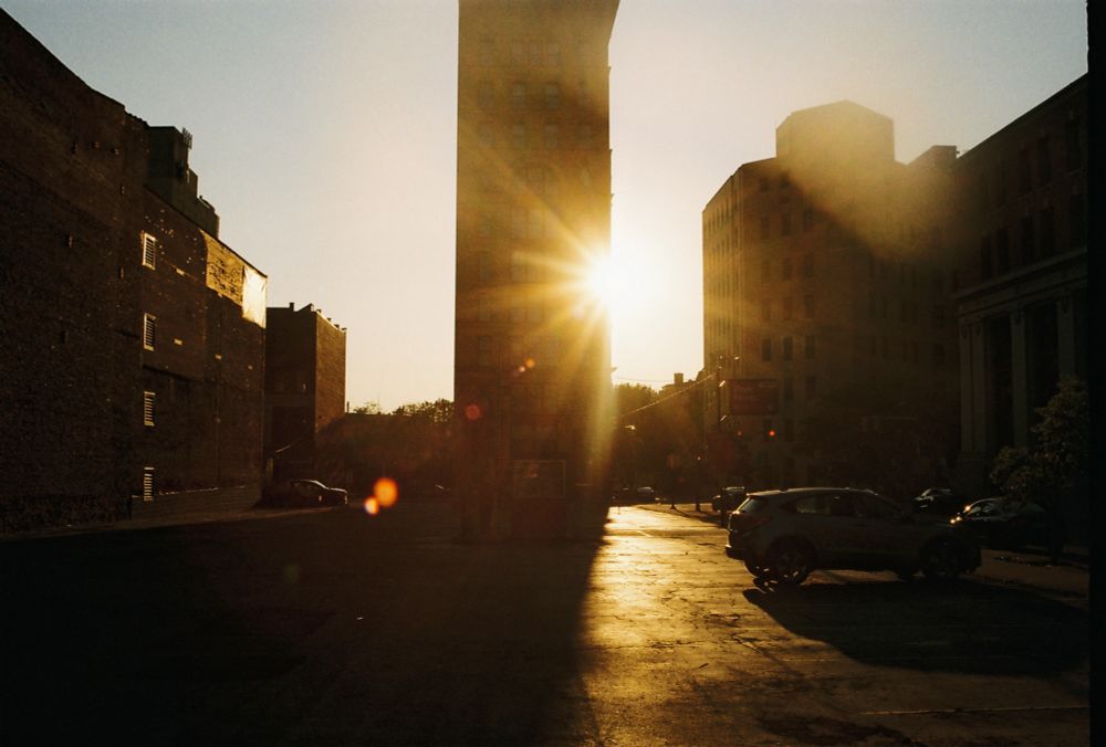 Sunset against several brick buildings in the city. There’s a parking lot and some cars strewn about. 