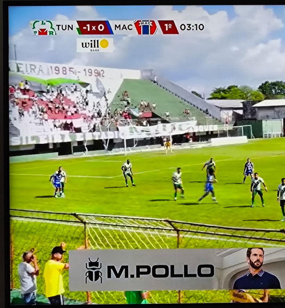 Foto de uma imagem de jogo de futebol reproduzido em uma Tv. Tuna luso x mac, o placar marca menos um pra tuna, zero par ao MAC