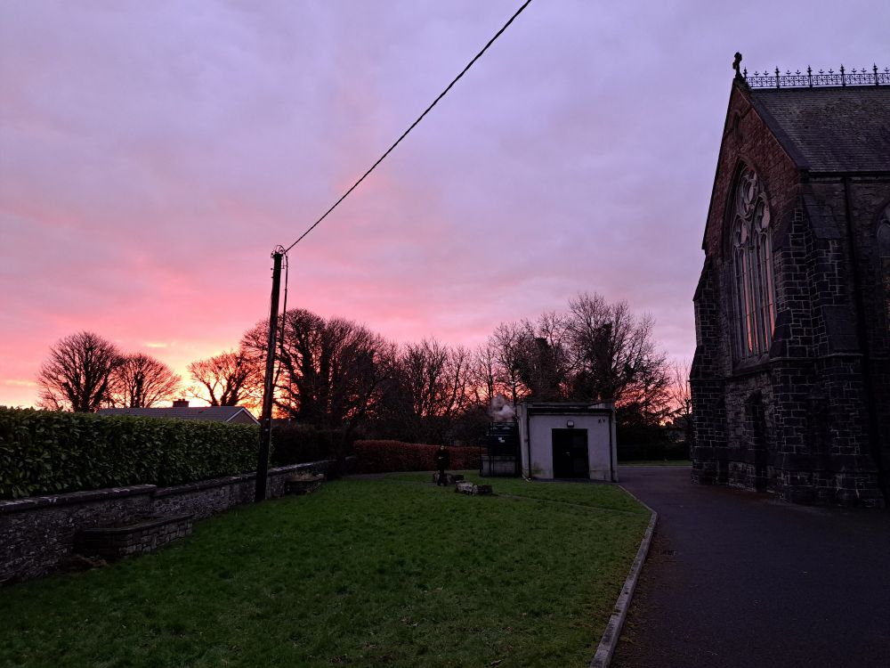 A photo of back of the church, sky is covered by clouds, a bright orange light of the sun is becoming visible 