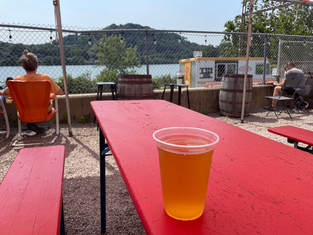 Photo of a pint of beer in a plastic cup looking out over a river and a green hill behind the river 