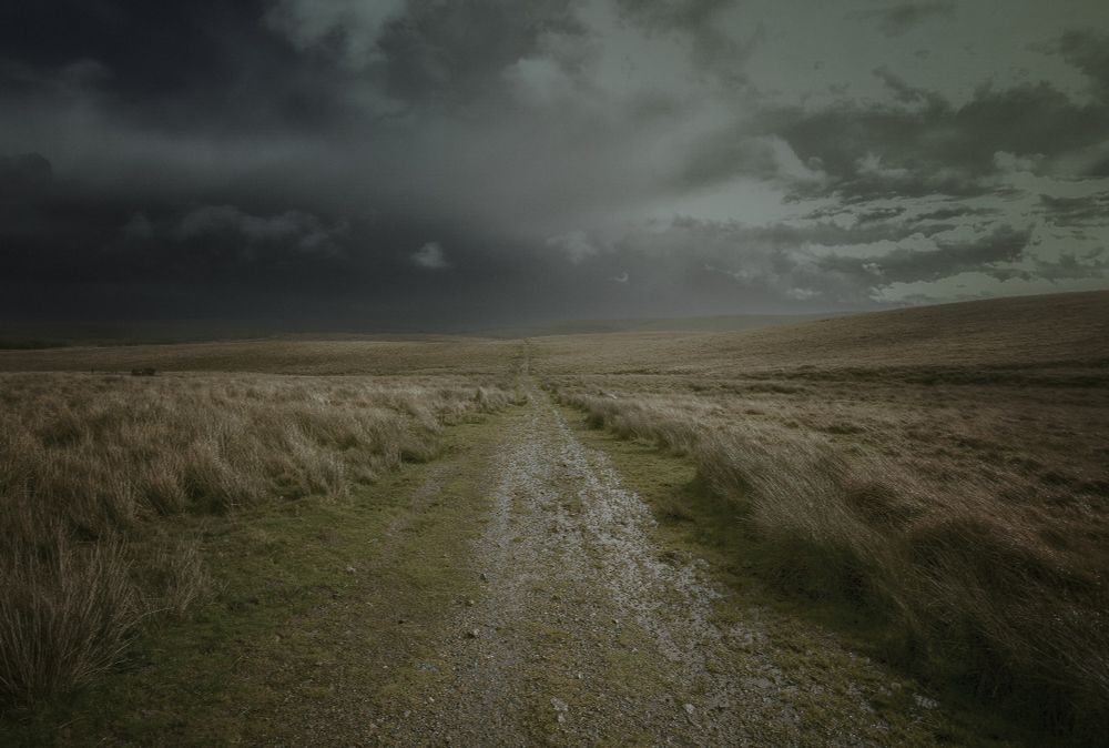 A narrow untamed road stretches into the distance across the Moor. Storm clouds gather on the horizon.