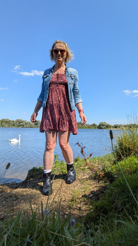 A woman in a sundress and denim jacket with chunky black boots standing beside a lake with a swan on it. It's bright sunshine and the sky is vibrant blue.