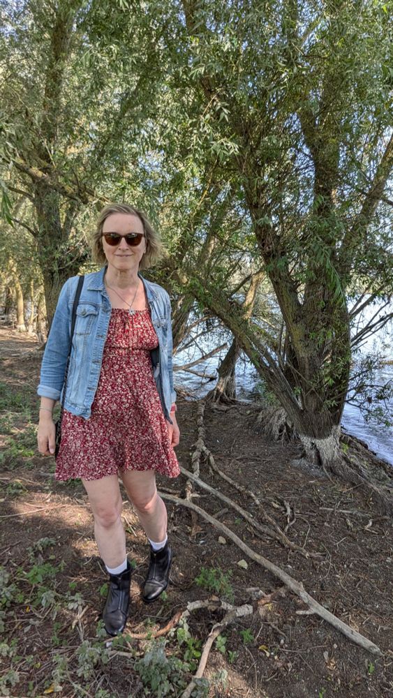 A woman with short blonde hair in a red ditzy sundress and blue denim jacket with black boots and sunglasses standing under a tree by a lake.