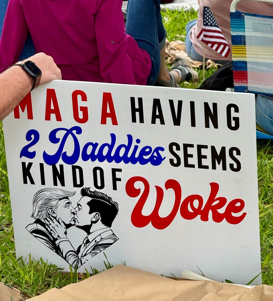 A protest sign on grass reads: “MAGA having 2 Daddies seems kind of Woke,” with colorful stylized text. Below, there’s a black-and-white drawing of Donald Trump and Elon Musk kissing,  and holding each other’s faces.