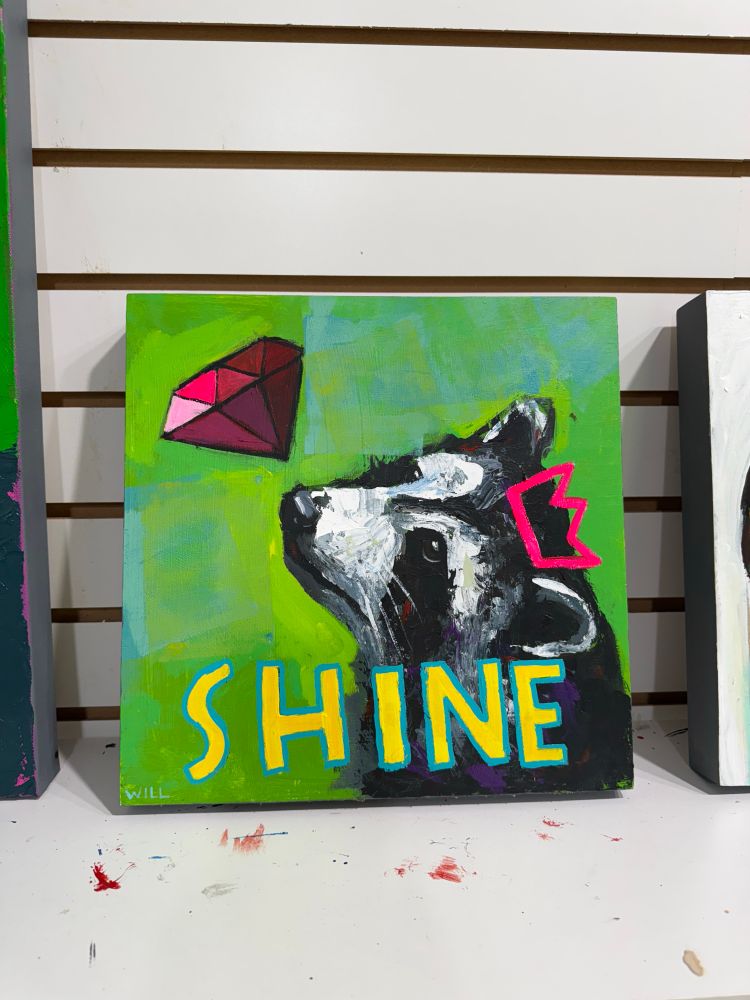 Colorful painting of raccoon by Will Eskridge with ruby gem and the words “Shine” in yellow letters. The raccoon is donning a crude pink king crown