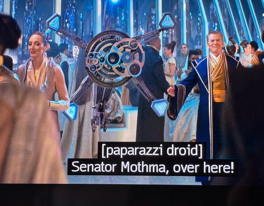 A paparazzi droid asking Mon Mothma to pose for a photo at a crowded, lavish ball.