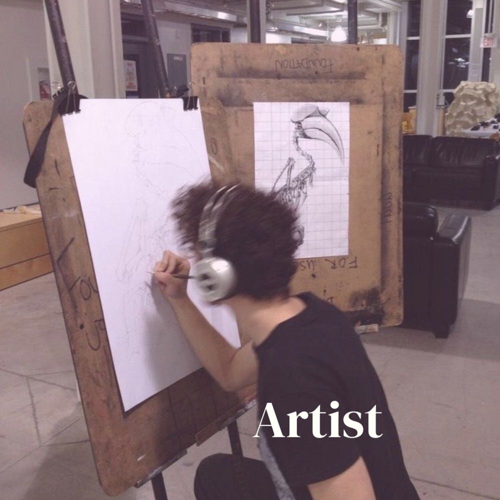 Foto de um homem branco de cabelo preto usando roupas pretas e um fone de ouvido cinza desenhando em uma folha que está em uma espécie de cavalete. Na foto, a palavra “artist” está escrita