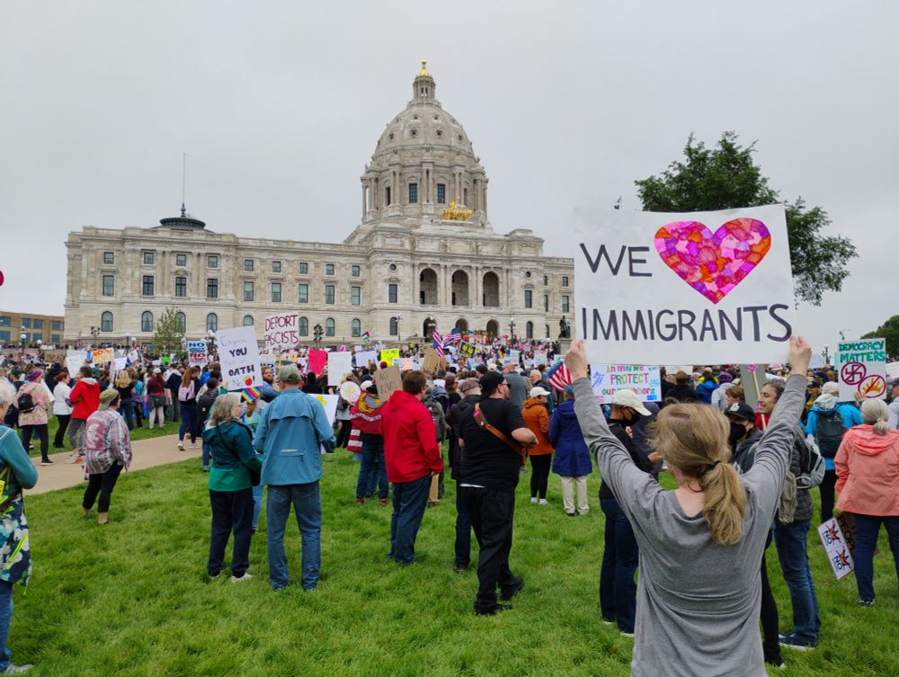 Scene of the Minnesota Capitol against a grey sky. A protester in the foreground is holding a sign that says "We Love Immigrants!" The word love is a heart drawn with a patchwork of multiple colors to represent solidarity.
