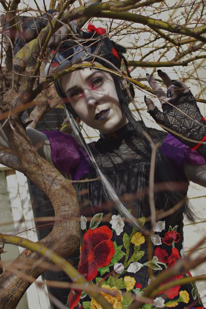 Kacie is dressed as Laudna from Critical Role in her level 9 outfit, stood behind branches in the foreground of the photo, some of them are blurry. She stands with her head slightly tilted staring at the camera and her left hand raised in a creepy way, the right holding onto the tree branch above their head. 
