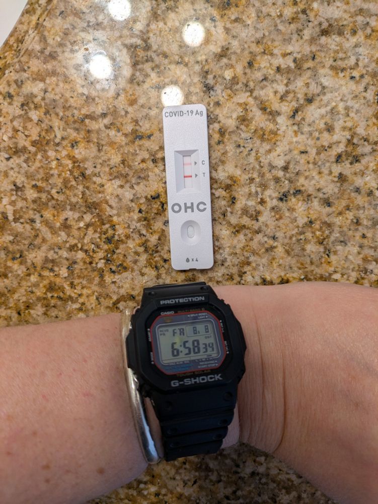 a photo of a positive covid test and a person's wrist next to it, wearing a digital watch and showing Friday, 8/8, and the time as 6:58