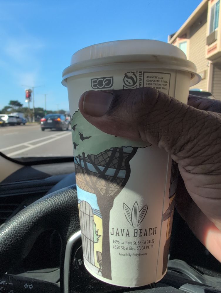 A man's hand holds a large coffee cup, partly filled with coffee from nearby Java Beach Cafe, with a plastic lid on it